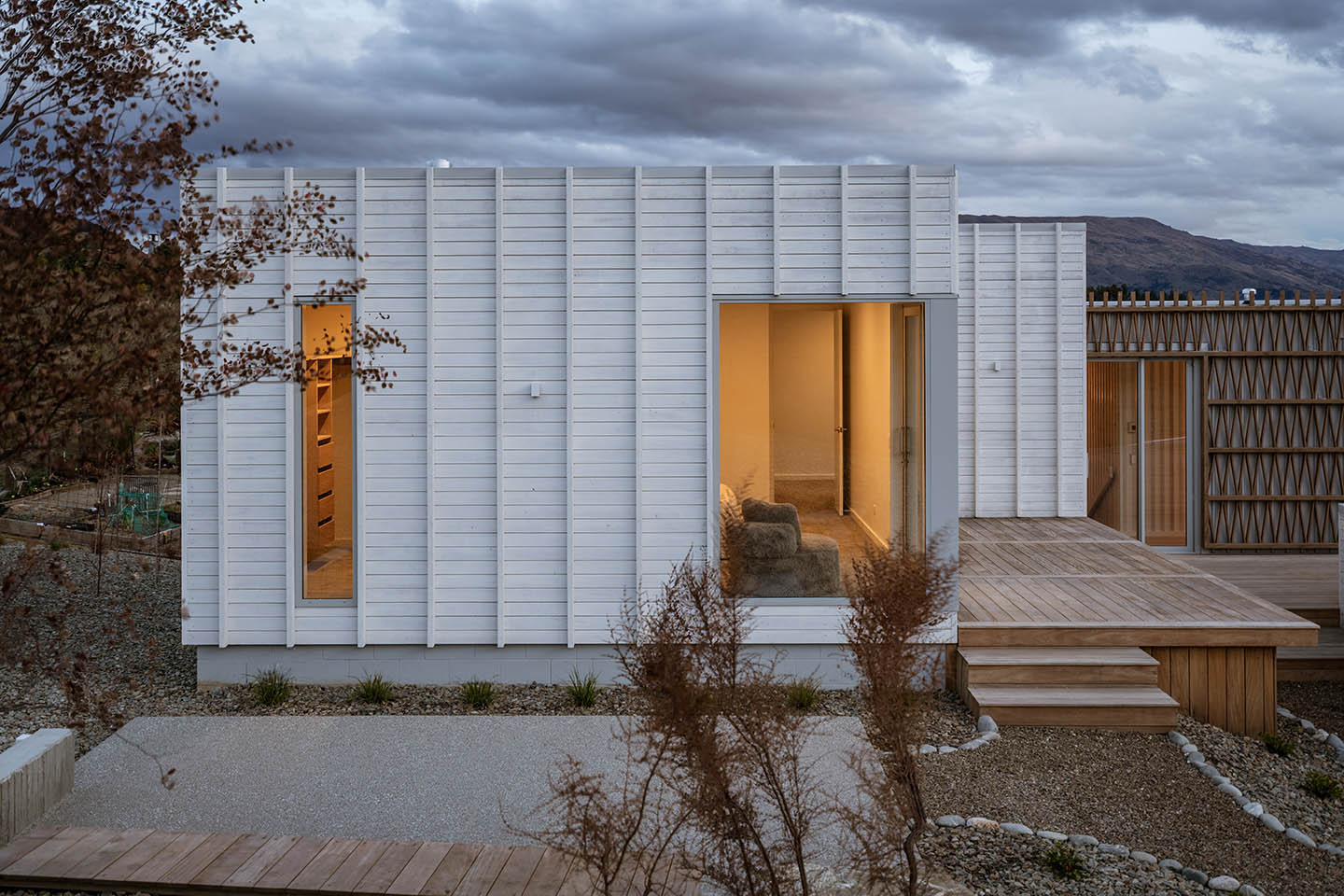 Akitu IV exterior at dusk with pale vertical cladding, narrow full-height windows and a timber deck connecting to the screened wing.