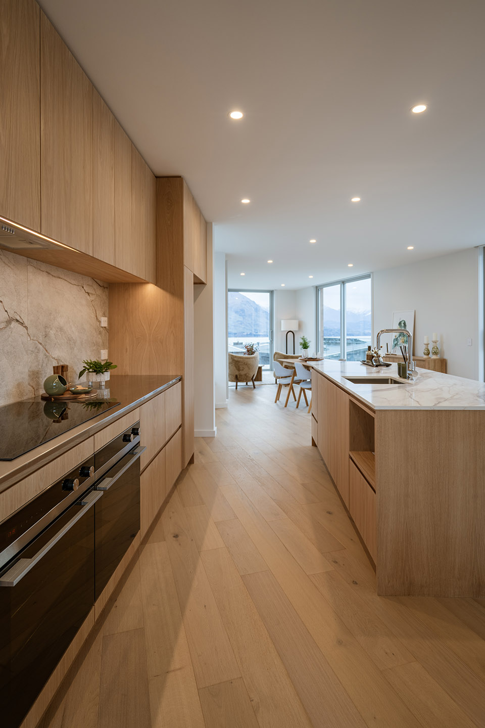 Akitu IV kitchen with timber cabinetry, pale stone surfaces and glazing beyond the dining area framing lake and mountain views.