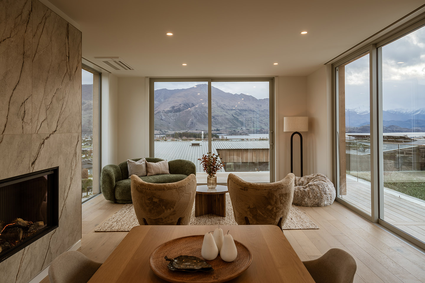 Akitu IV living area with a stone-lined fireplace and full-height glazing framing Lake Wānaka and the surrounding mountains.