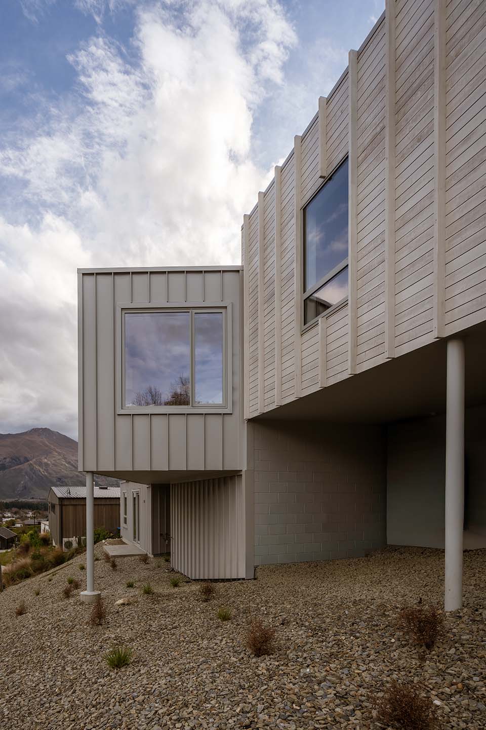 Akitu IV exterior showing pale vertical cladding, large framed windows and upper forms raised above the sloping landscaped ground.
