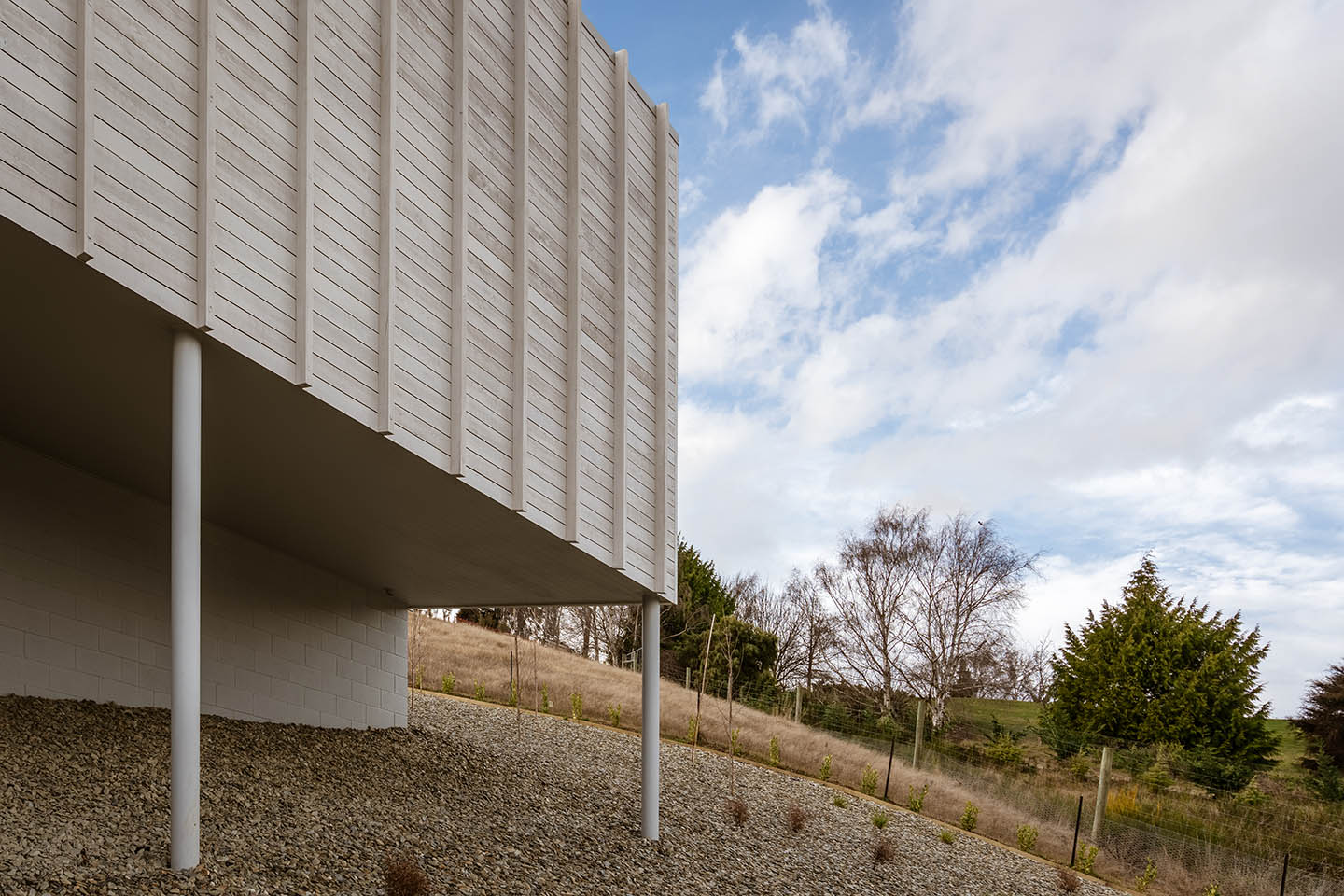 Akitu IV exterior detail showing the pale upper volume supported above the sloping gravel landscape on slender columns.