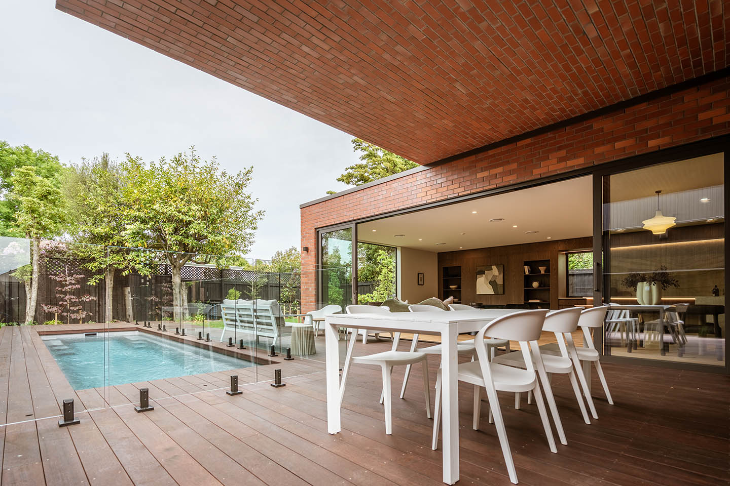 Bryndwr II pool courtyard with outdoor dining beneath the brick overhang and wide glazing opening into the living spaces.