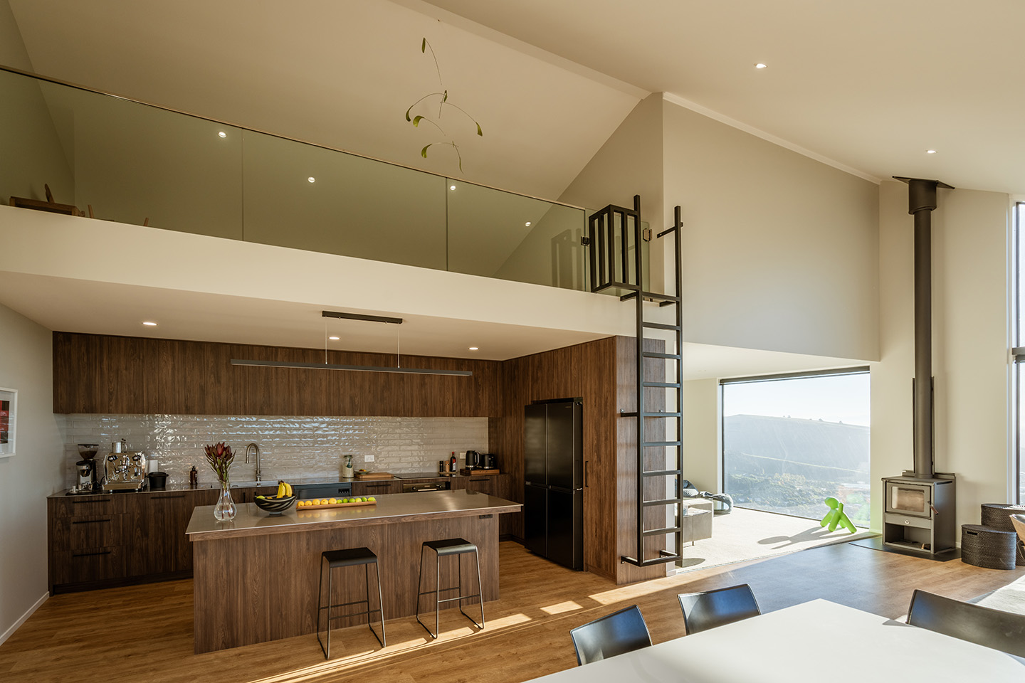 Major Hornbrook II kitchen with timber cabinetry and island beneath the loft, beside a freestanding fireplace and tall hillside glazing.