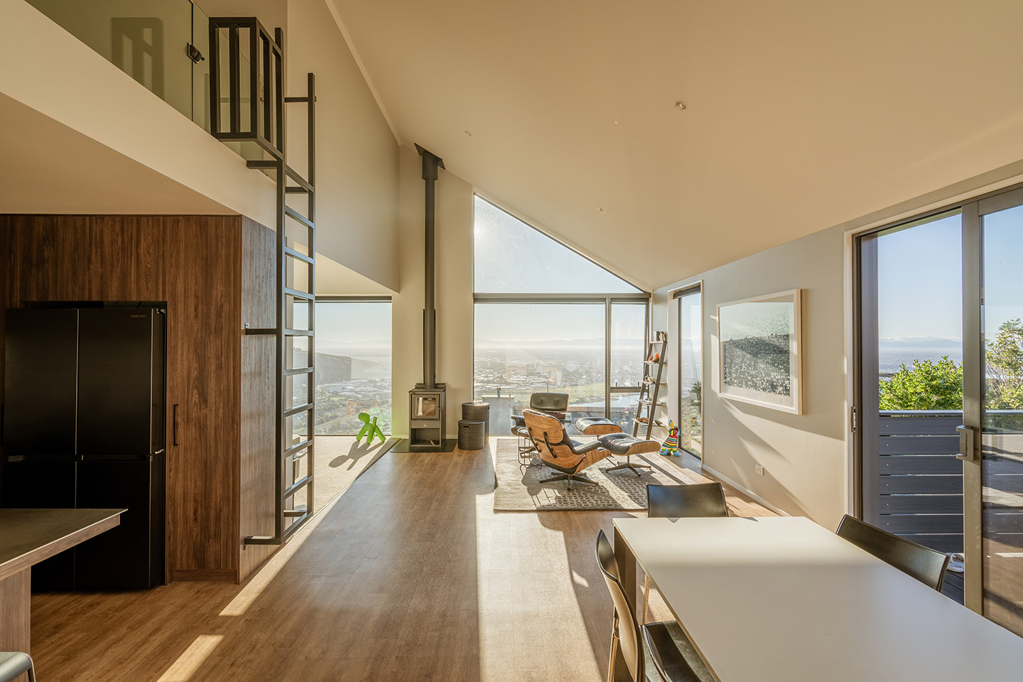 Major Hornbrook II living and dining area beneath a rising ceiling, with a steel loft ladder and full-height glazing toward the view.