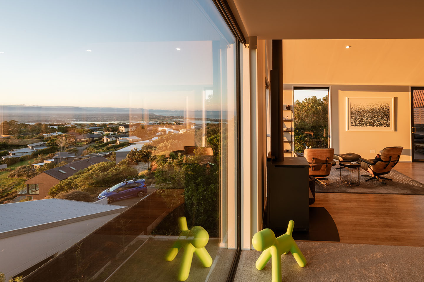 Major Hornbrook II living area beside floor-to-ceiling glazing, with the hillside neighbourhood, estuary and coastline visible below.