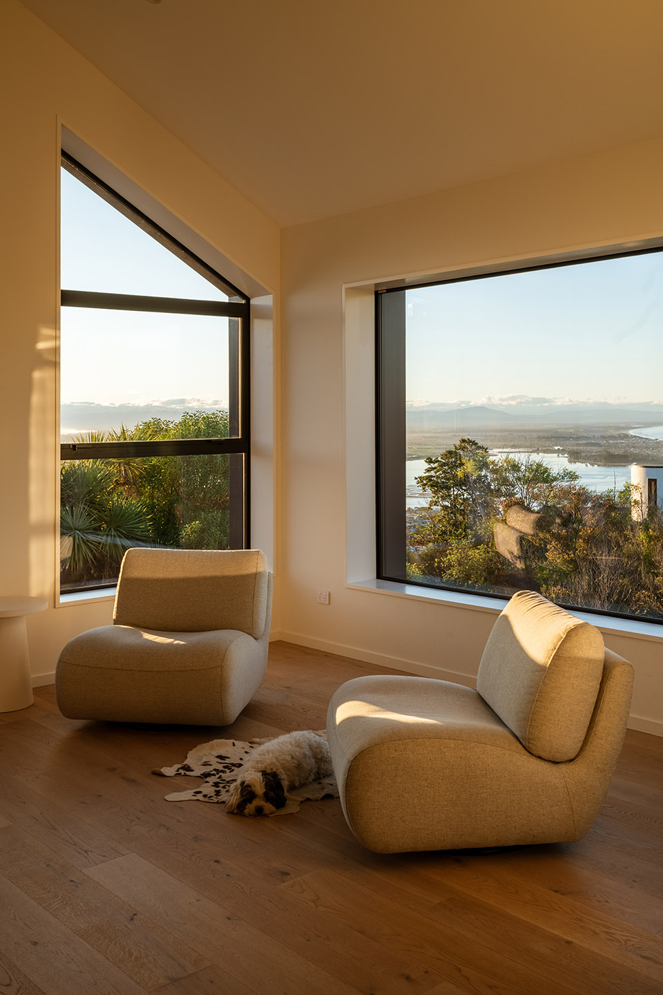 Revelation III sitting area with timber flooring and two large windows framing the estuary, coastline and surrounding vegetation.